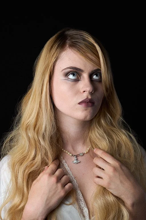 Portrait of a blonde woman against a dark background looking upward with a neutral expression, wearing a pearl necklace with a silver pendant and a white lace top. Fashion