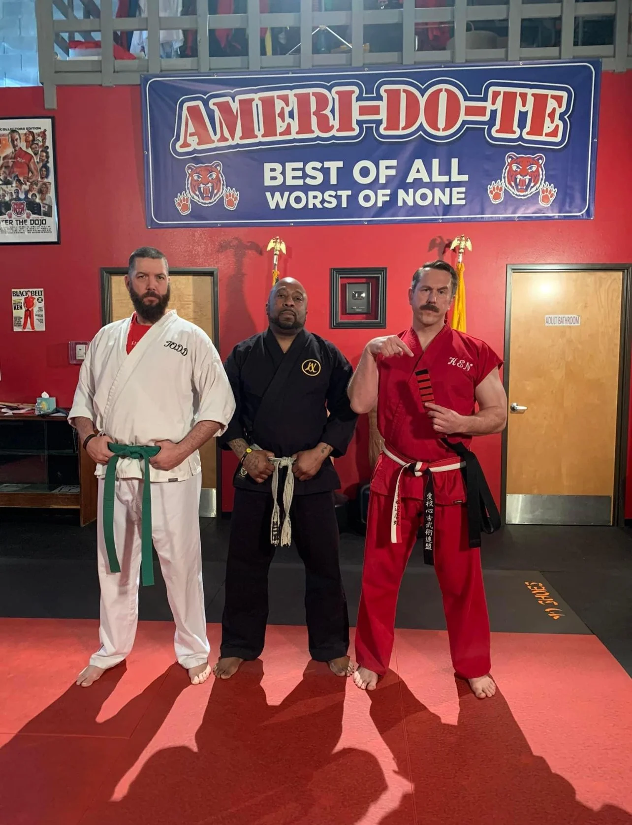Three men in martial arts uniforms standing on a red mat in a dojo. The wall behind them has a large sign saying 'AMERI-DO-TE - Best of All, Worst of None,' with a bear mascot. There are flags and a door to the adult bathroom on the right.