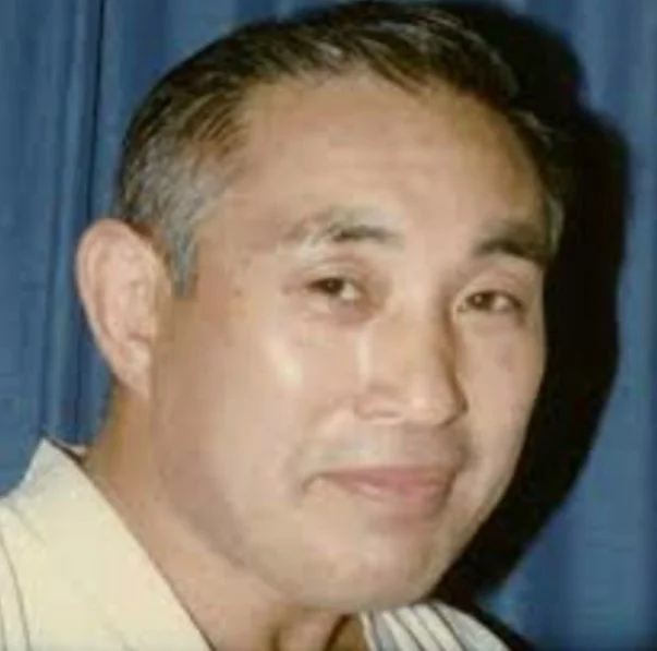 A man with short dark hair and light skin, smiling slightly, standing in front of blue curtains.