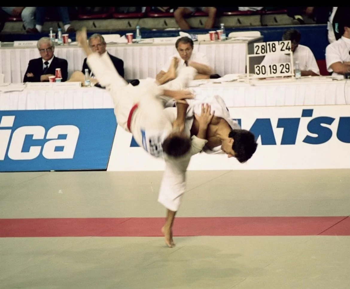 Two judo athletes engaged in a match, with one executing a throw on the other. Several judges or officials sit behind a table in the background, observing the match, with a scoreboard displaying scores.