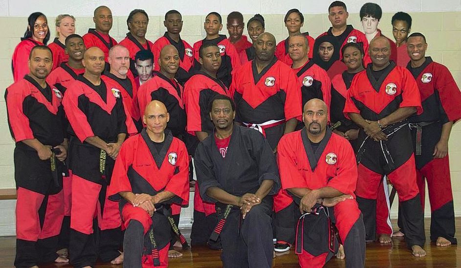 Group of martial arts practitioners in red and black uniforms, some with black belts, posing for a photo indoors.