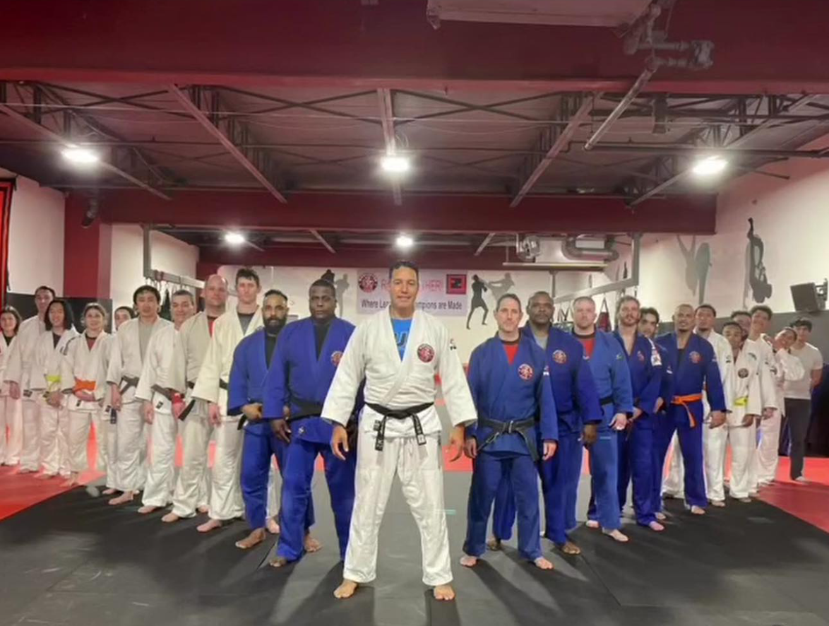 Group of martial artists in gi uniforms, standing in formation inside a gym, with some wearing black belts and others colored belts, under a red ceiling with martial arts silhouettes on the walls.