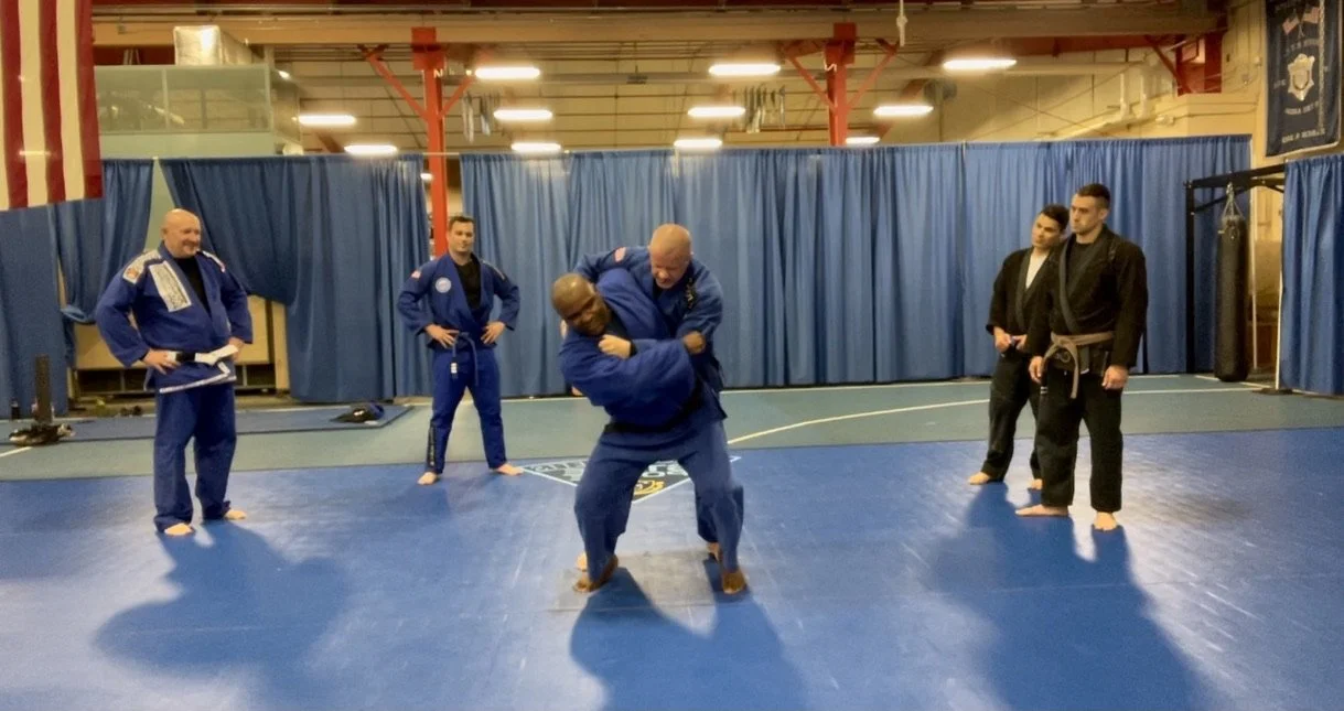 Two martial artists practicing grappling or Brazilian Jiu-Jitsu on a blue mat in a training facility with onlookers, some in gi and some in casual clothing.