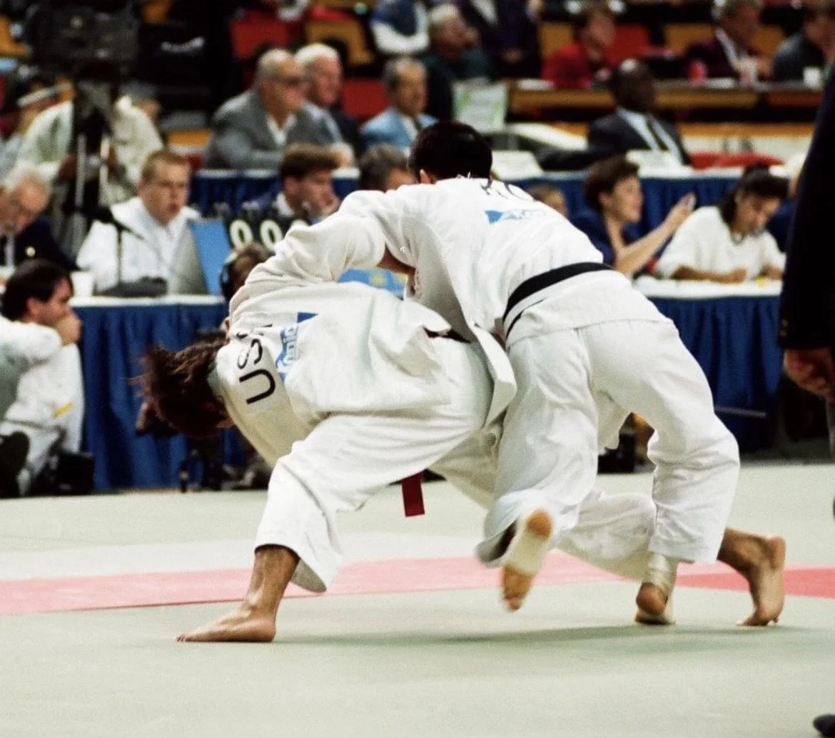 Two judo competitors in white gis battling on the mat during a match at a tournament, with spectators watching in the background.