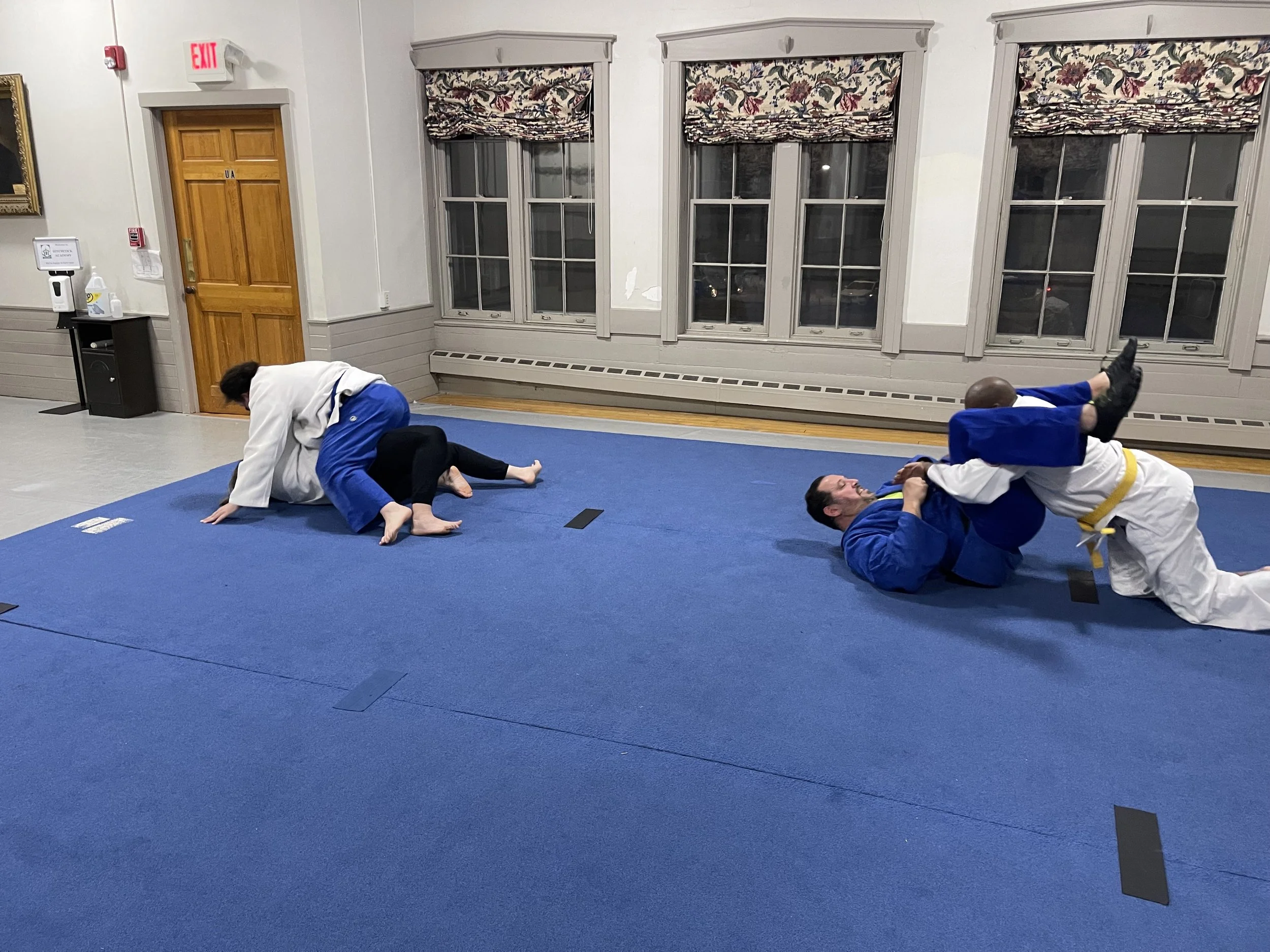 Two pairs of people practicing Brazilian Jiu-Jitsu on a blue mat in a room with wooden doors and large windows. One pair is engaged in a ground grappling position, and the other pair is on the floor, with one person in a white gi and yellow belt.