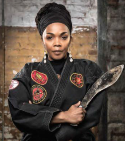 A woman wearing a black traditional outfit and head wrap, holding a large curved knife and standing in front of a brick wall.