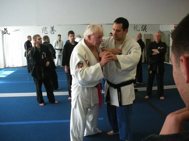 Two martial artists practicing a technique in a dojo with other students observing.