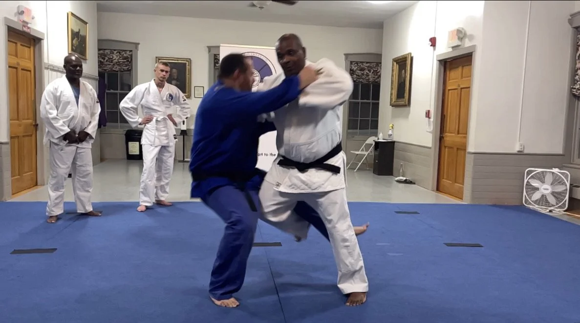 Two men practicing Brazilian Jiu-Jitsu in a dojo while three other men watch.
