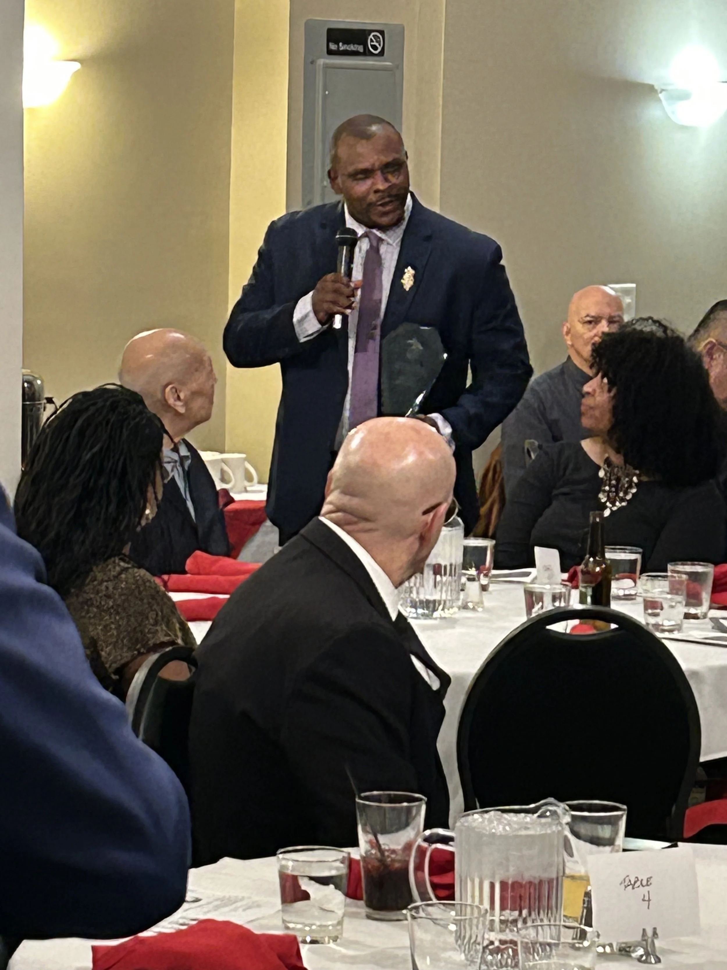 A man in a dark suit and purple tie speaking into a microphone at a banquet or conference, with seated attendees listening around the table.