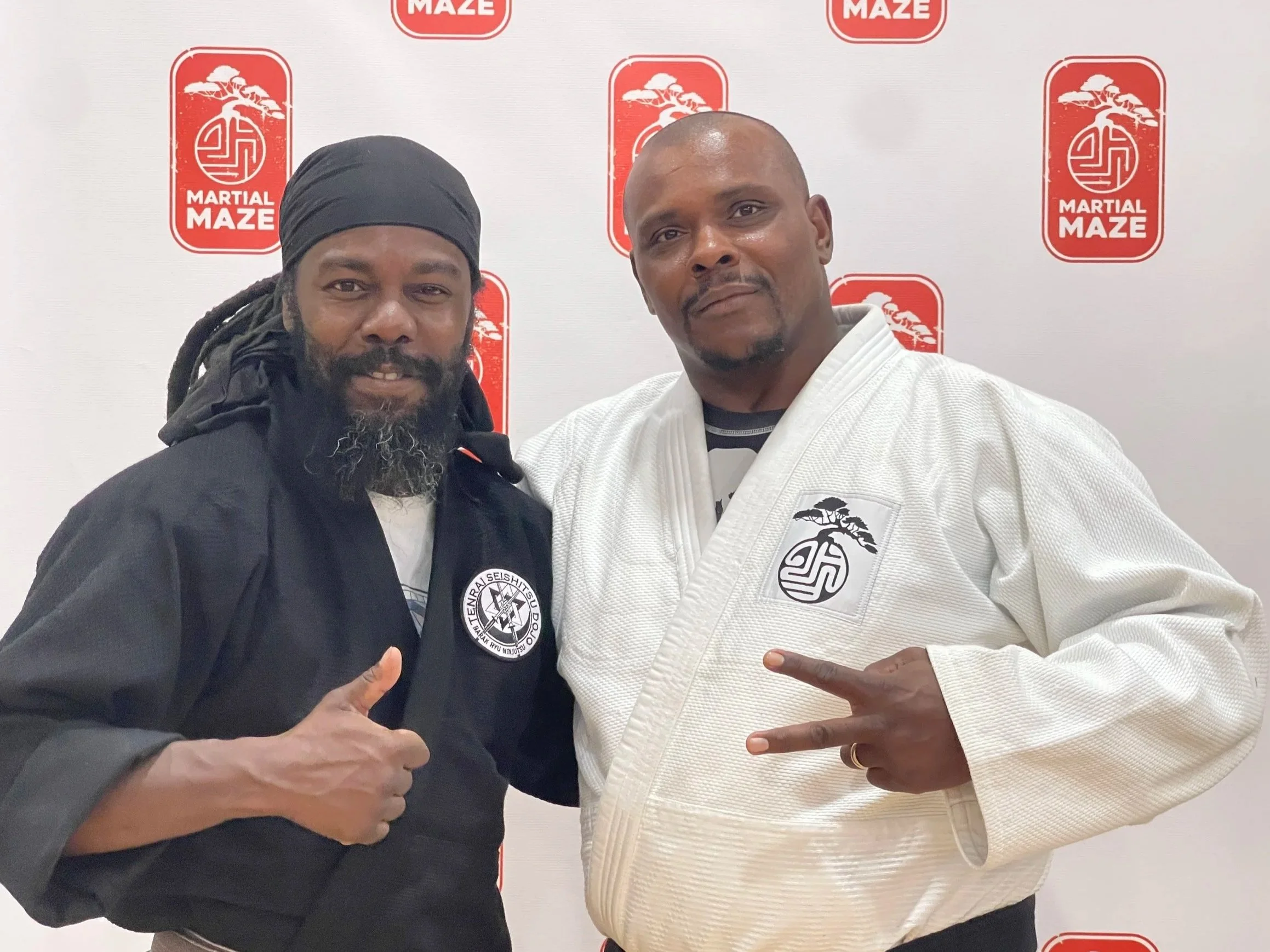Two men in martial arts uniforms posing in front of a backdrop with the 'Martial Maze' logo. The man on the left is wearing a black uniform and giving a thumbs-up, and the man on the right is wearing a white uniform, making a peace sign with his fingers.