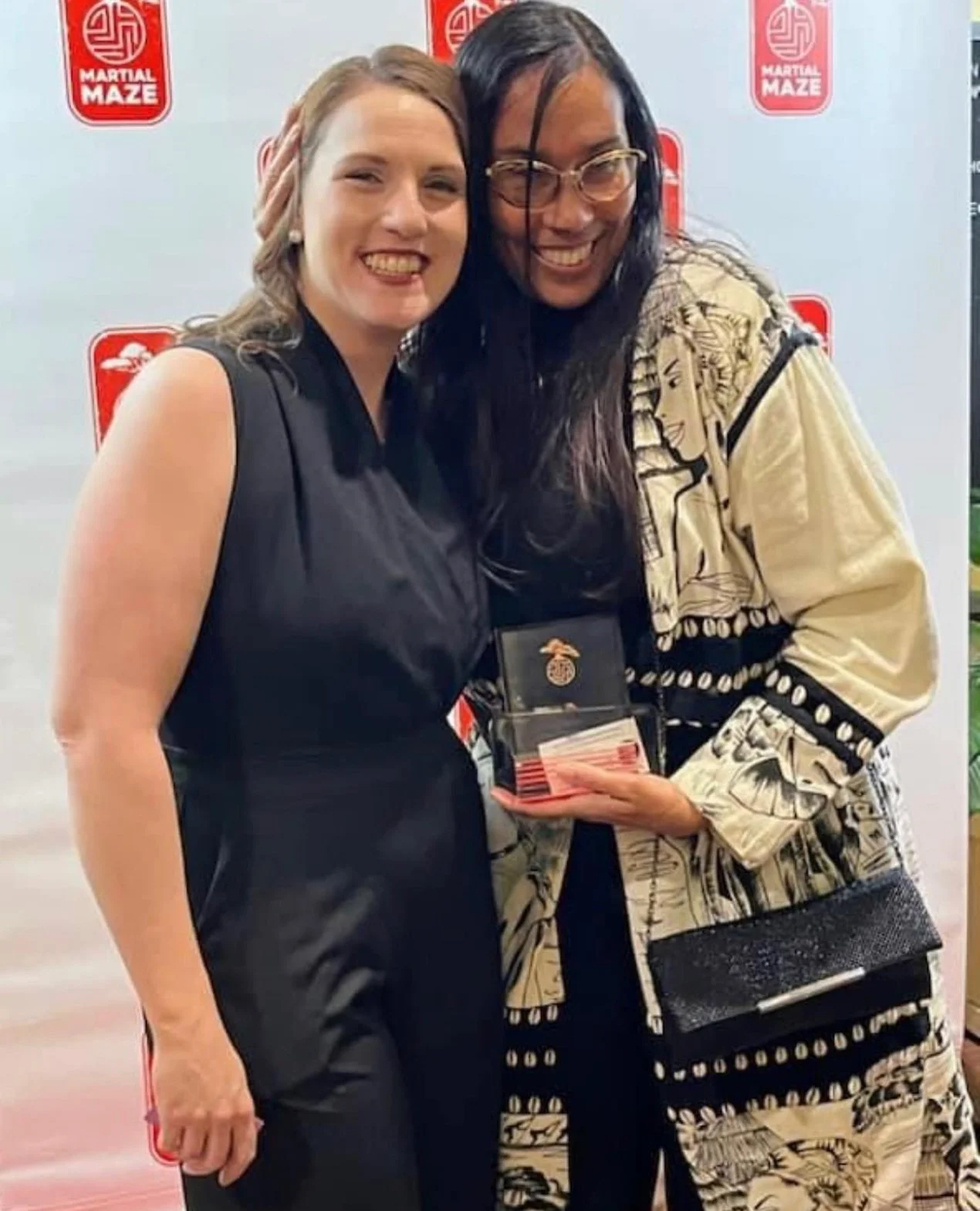 Two women smiling, one holding a medal and certificate in front of a 'Martial Maze' backdrop.