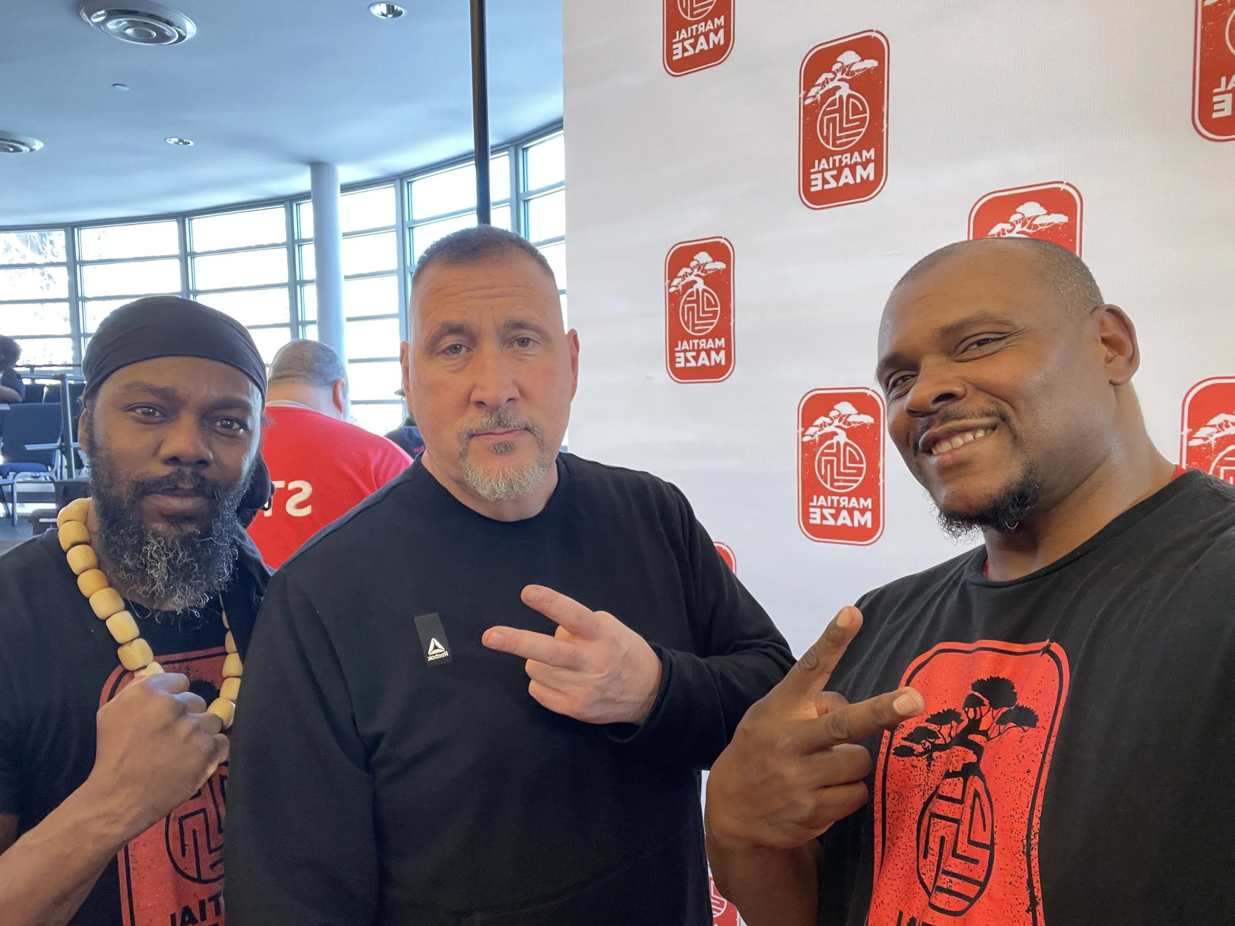 Three men posing for a photo at an event with a Machail Maze logo in the background. The man on the left wears a black headband and necklace, the man in the middle has a beard and makes a peace sign, and the man on the right smiles and points at the camera.