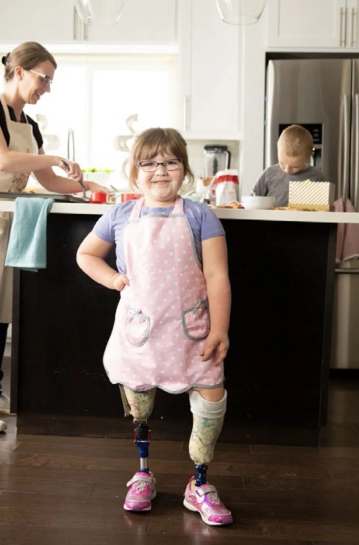 A young girl with a prosthetic leg standing in a kitchen, wearing a pink apron over a blue shirt and pink sneakers, smiling at the camera. In the background, a woman and a boy are preparing food.