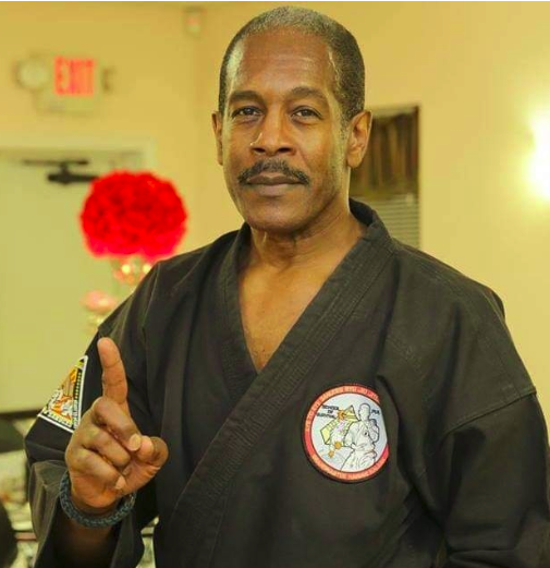 A man in a martial arts uniform poses indoors, holding up one finger, with a red flower decoration and an exit sign visible in the background.