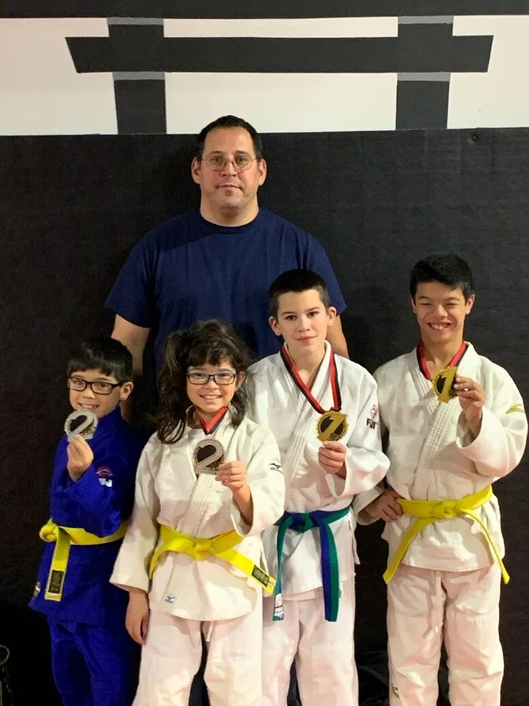 Group photo of four children in judo uniforms with medals and an adult man standing behind them, in an indoor martial arts gym.