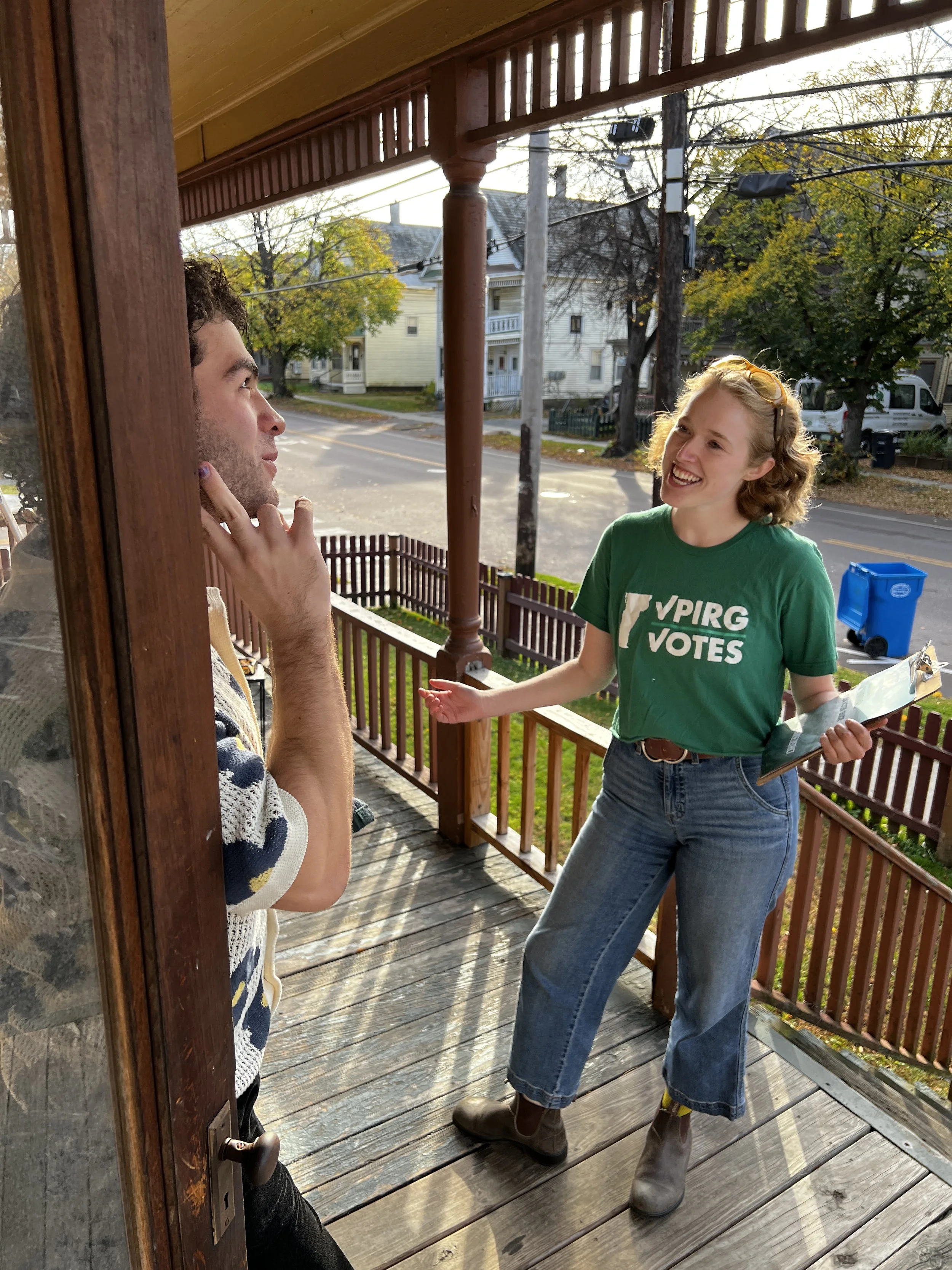 A canvasser wearing a VPIRG Votes T-shirt holding a clipboard, having a conversation with a man on a sunlit porch.