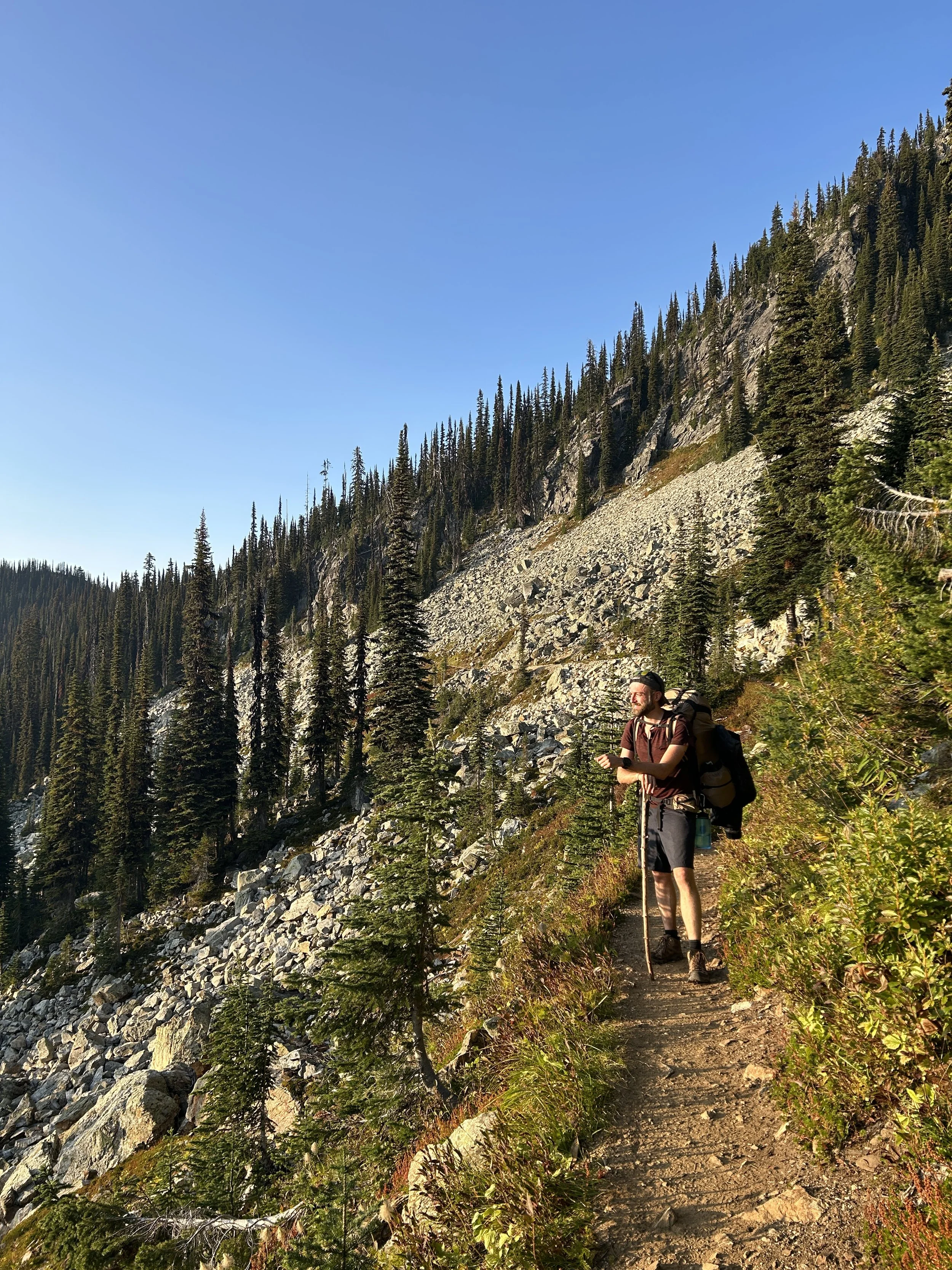 A man hiking on a narrow trail in a mountainous forested area with tall evergreen trees and rocky slopes.