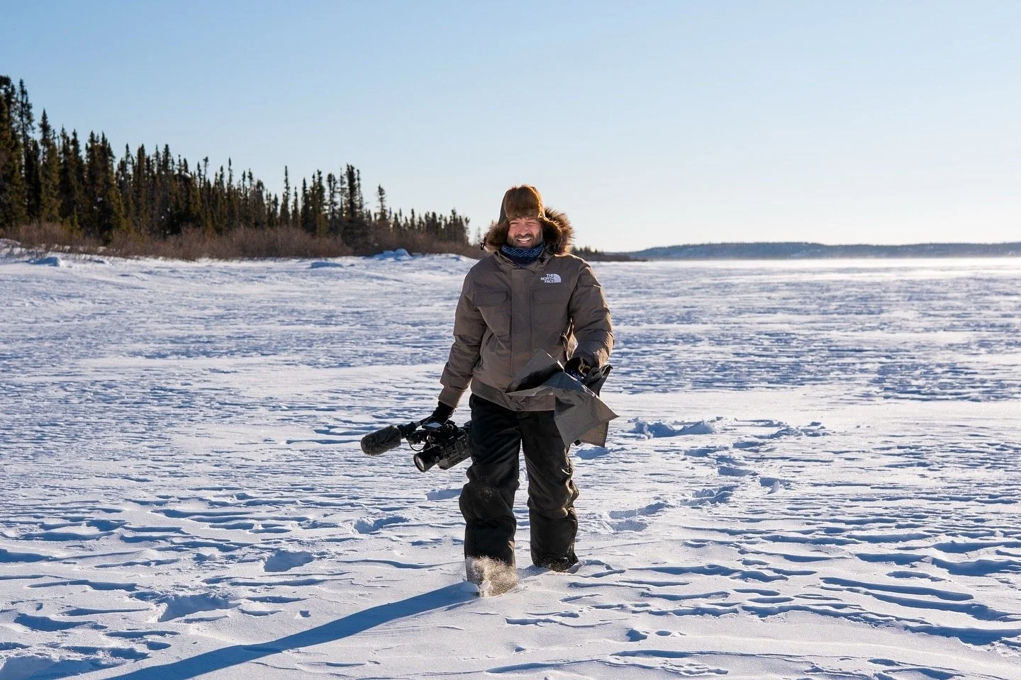 Person in winter clothing walking on snow-covered landscape with trees in the background, holding a camera and some equipment.