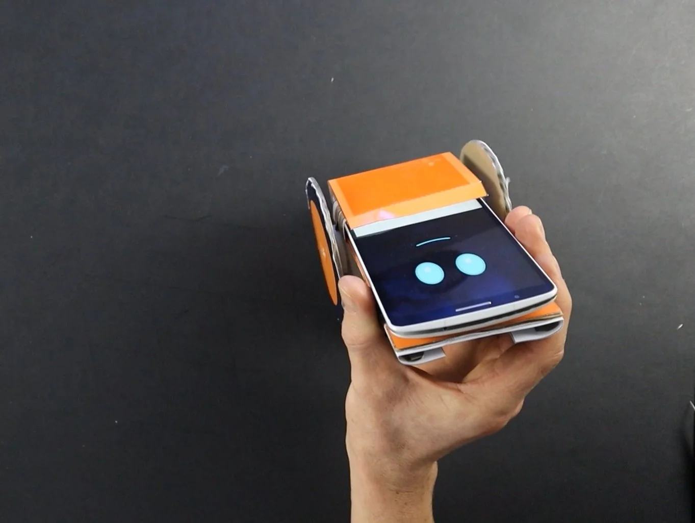 smartphone robot held by a hand ready to start. the smartphone robot is orange with the smartphone showing a blue smiley face