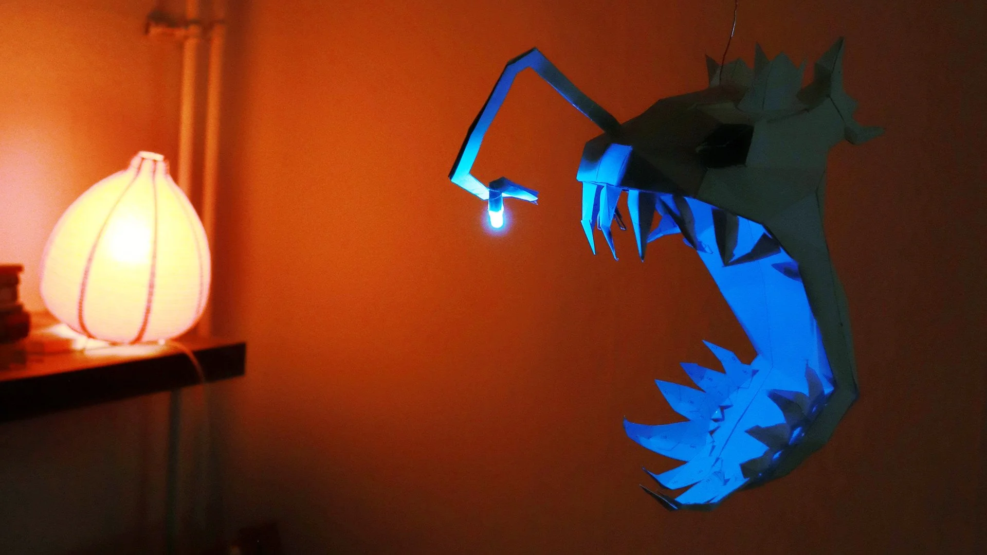 A low poly angler fish made from paper. The angle is light by an color-changing LED (light blue). The background is illuminated by a red lamp giving the picture a red-blue contrast.