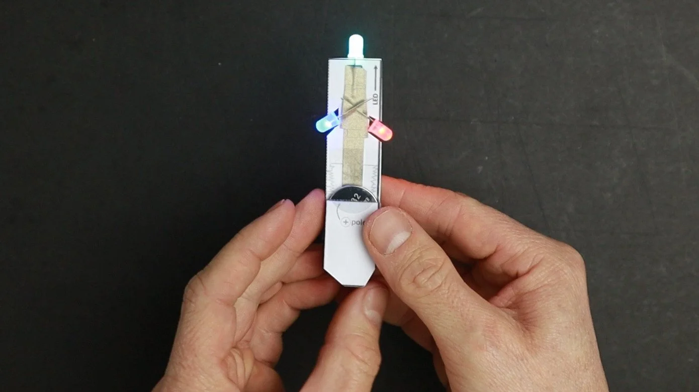 Two hands holding the finished paper circuit that connects a button cell to three rgb LEDs. The circuit is just a strip of folded paper with the LEDs on top and the button cell at the bottom.