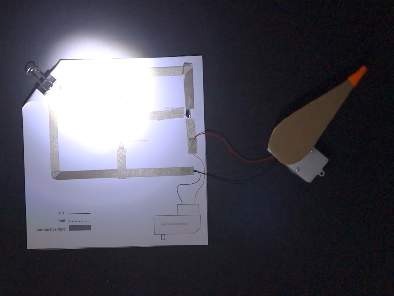Picture of the light-sensing circuit conneced to a geared motor with a large arrow on top to show the movement. the circuit is light with a bright spot and the arrow on the motor is turning.