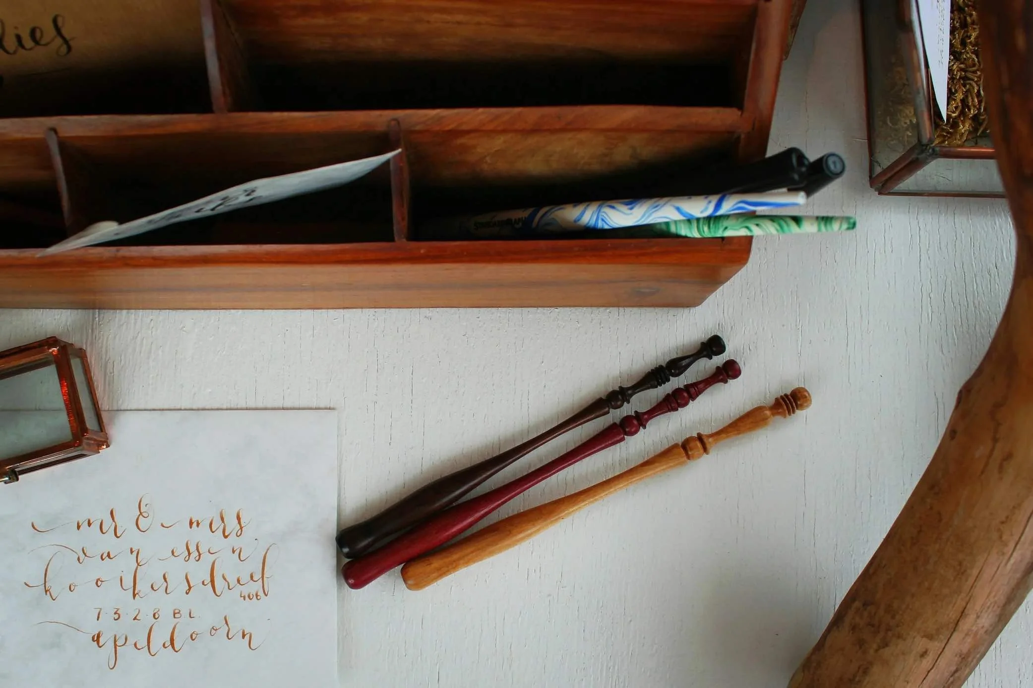 A wooden organizer with several compartments, containing a walking cane, a toy sword, a marbled pen, and a rectangular candleholder, placed on a white surface next to handwritten calligraphy paper.