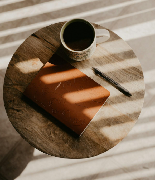 A wooden round table with a coffee mug, a black pen, and a closed orange notebook on it, with sunlight casting shadows through window blinds.