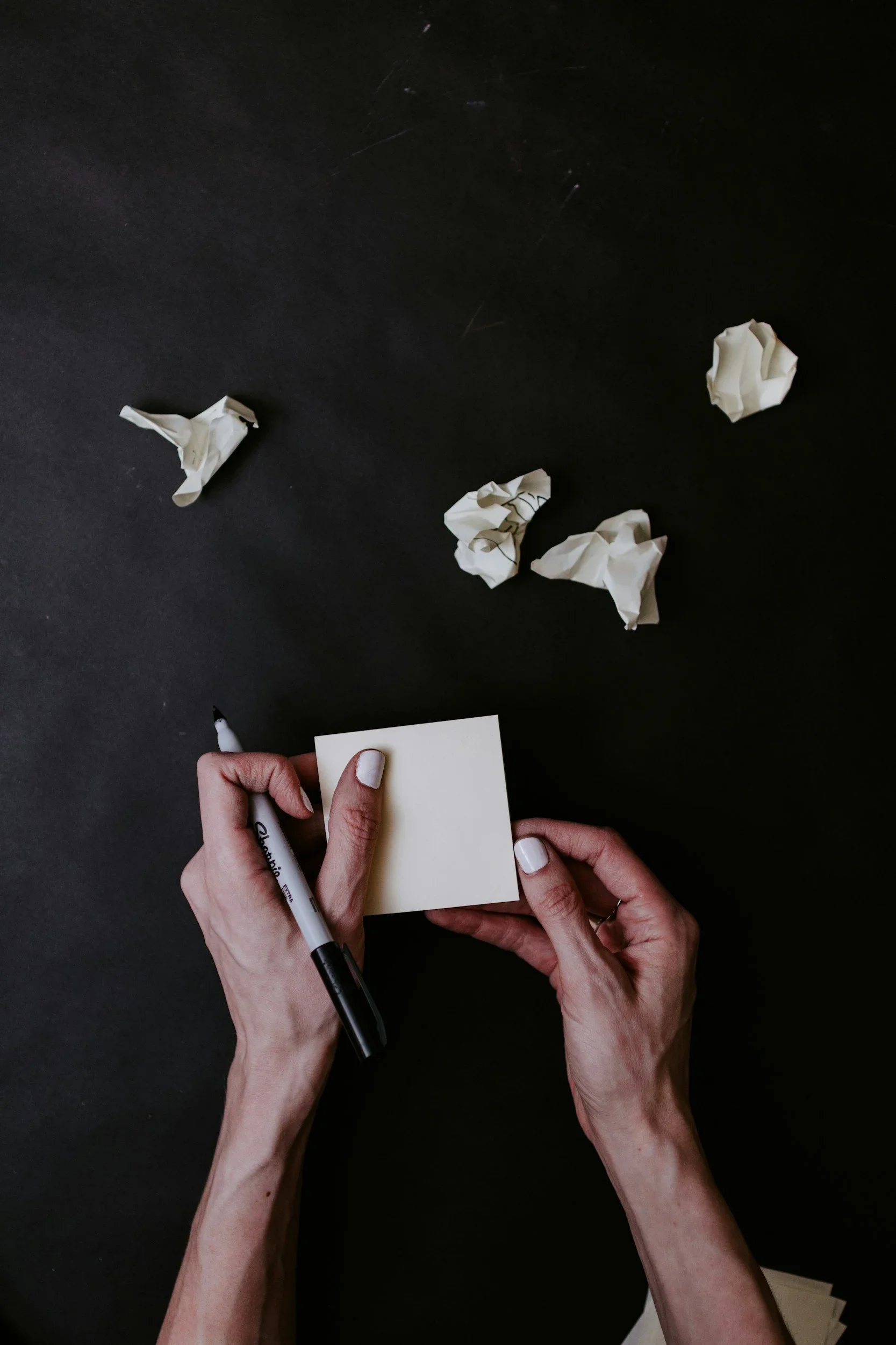 A person holding a sticky note and a black marker, with several crumpled pieces of paper on a dark surface and a hand writing on the sticky note.