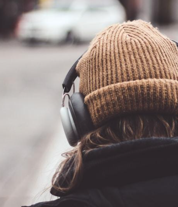 Close-up of a person wearing a brown knit beanie and black headphones, with their back facing the camera, outdoors.