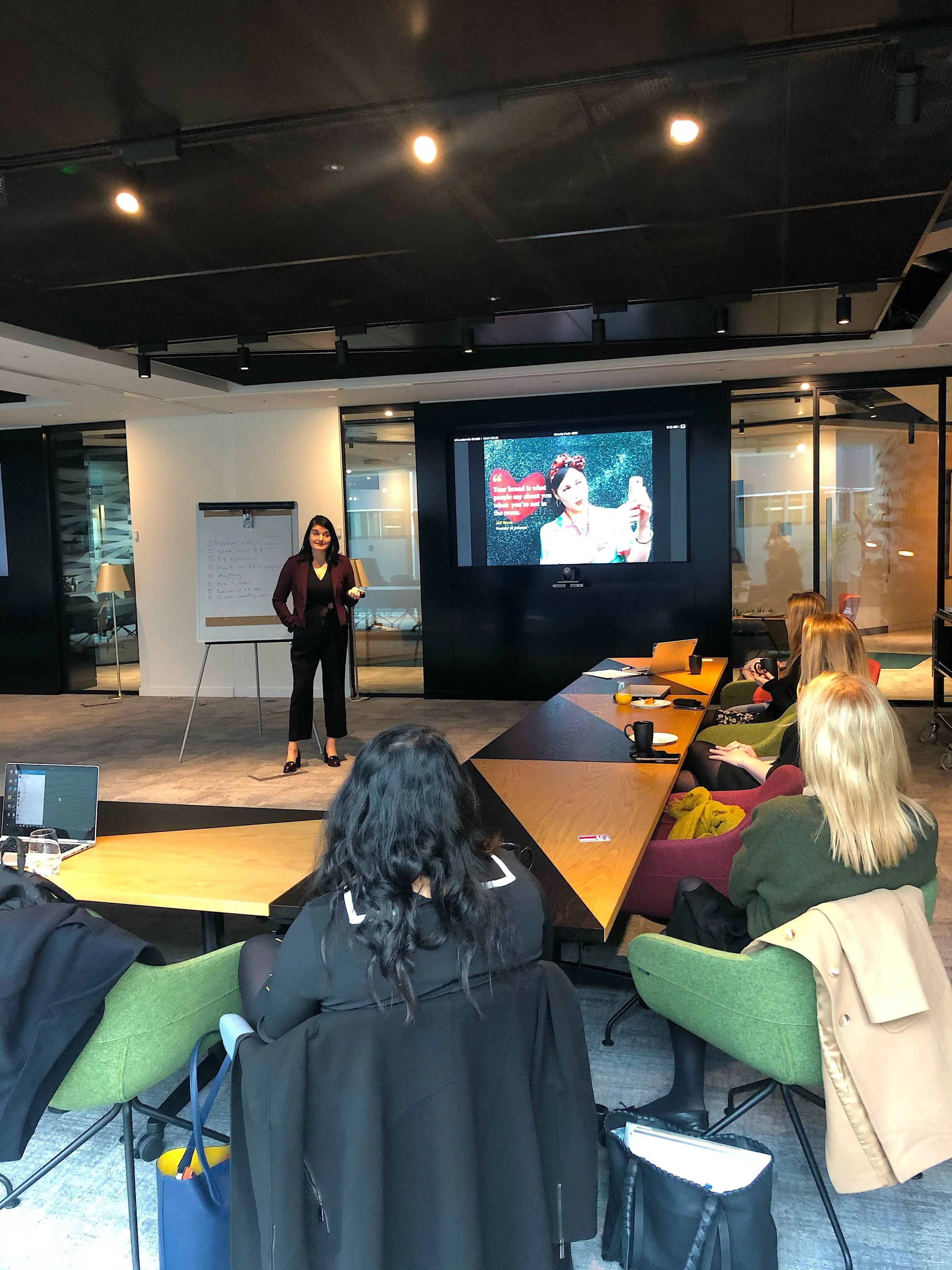 A woman (Monique Shaw) giving a presentation to a group of women in a modern conference room. The presenter stands near a large screen displaying an image of a woman taking a selfie, and there is a whiteboard with writing on it nearby.