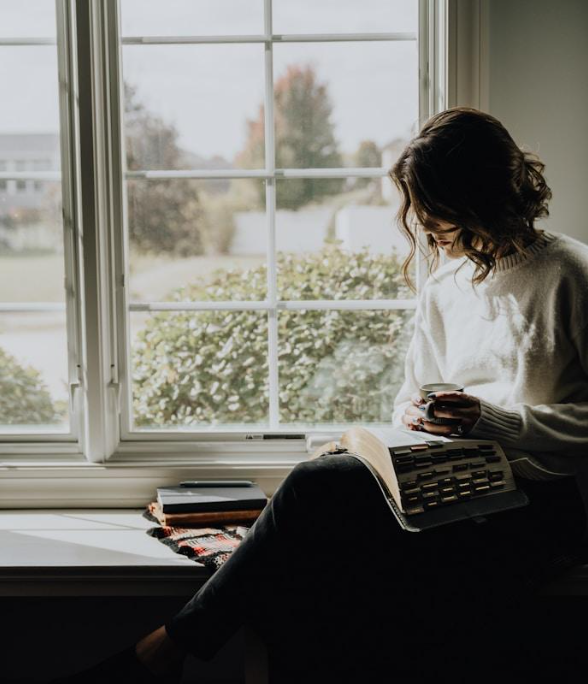 A woman sitting by a large window, looking at her phone, with a book on her lap, overlooking a garden with bushes and trees.