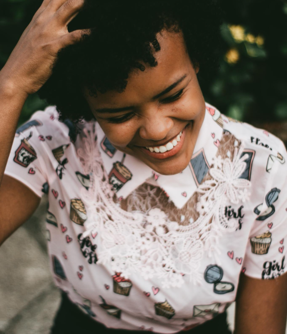 Smiling woman with curly hair wearing a colorful shirt with cupcake and makeup patterns, adjusting her hair outdoors.