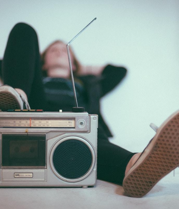 Person lying back with feet up, wearing sneakers, next to a vintage boombox, on a flat surface against a plain wall.