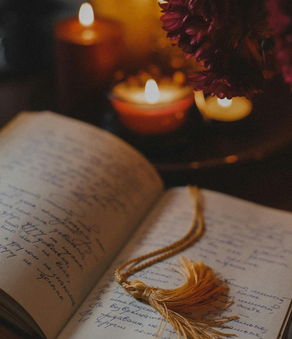 Open diary with handwritten text and a golden tassel bookmark, illuminated by candles and next to a bouquet of dark purple flowers.