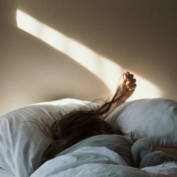 A person sleeping in bed with dark hair, partially covered by white bedding, with their arm extended and hand resting on a pillow. Sunlight creates a bright diagonal streak across the wall.