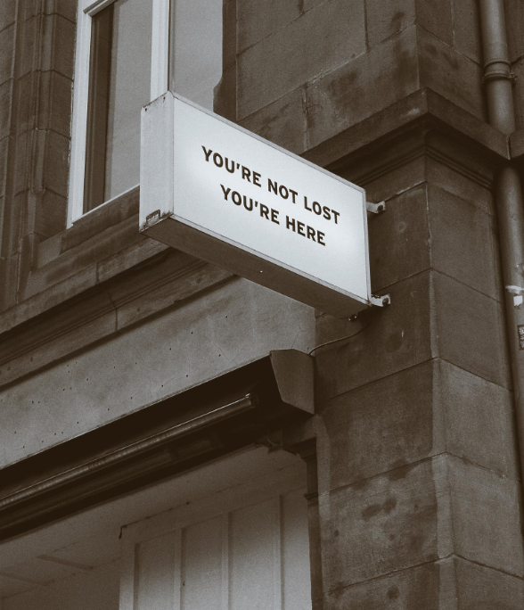 A rectangular white sign with black text that says "You're not lost, you're here" is mounted on the corner of a stone building.