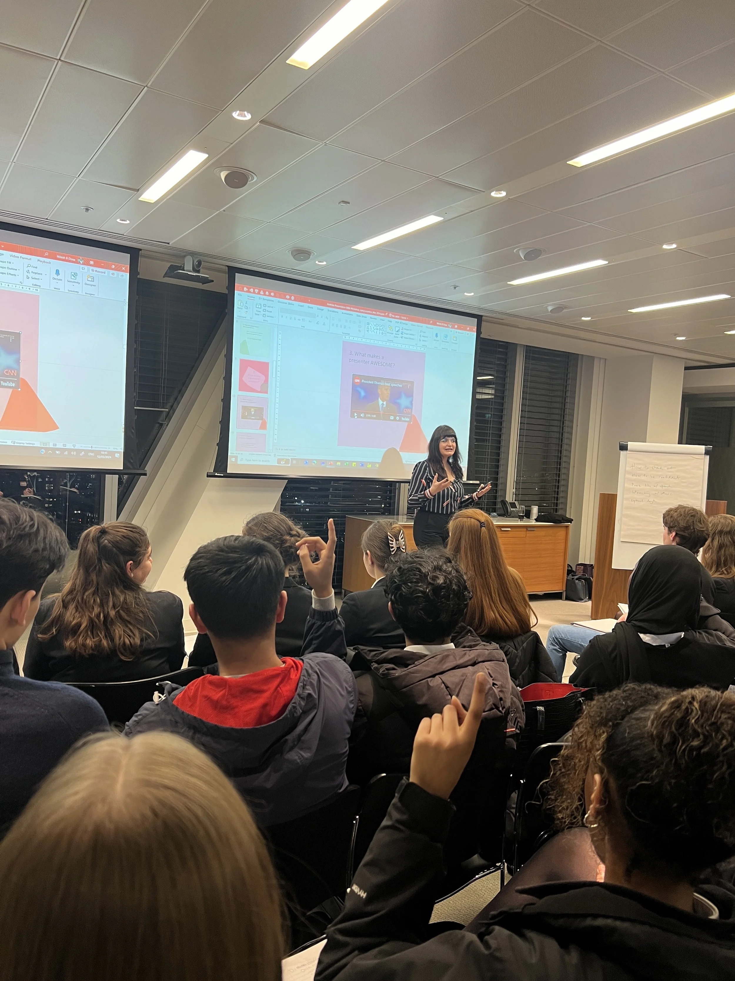 A woman is standing at the front of a room giving a presentation to a group of people seated in chairs. Two large screens display slides with text and images. The room has a high ceiling with lights and large windows with blinds.