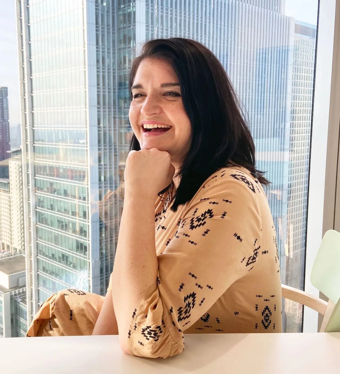 A woman with dark hair smiling and resting her chin on her hand, sitting at a table in front of a large window with a cityscape of tall skyscrapers in the background.