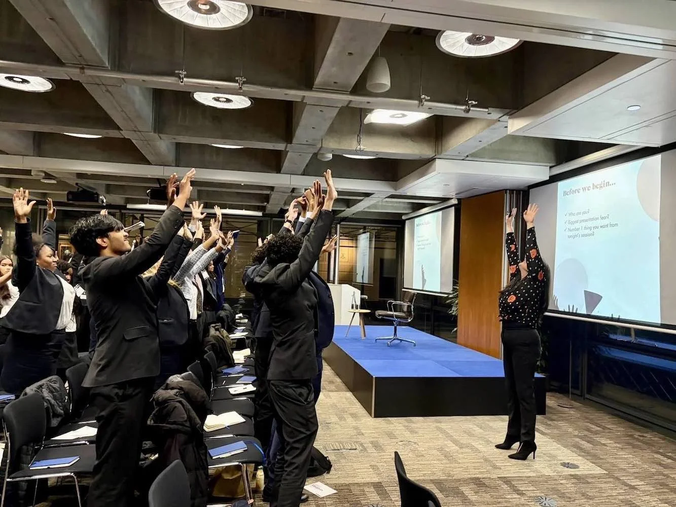 A group of people standing in a conference room, raising their hands during a presentation. A woman at the front stands near a large screen with a slide.