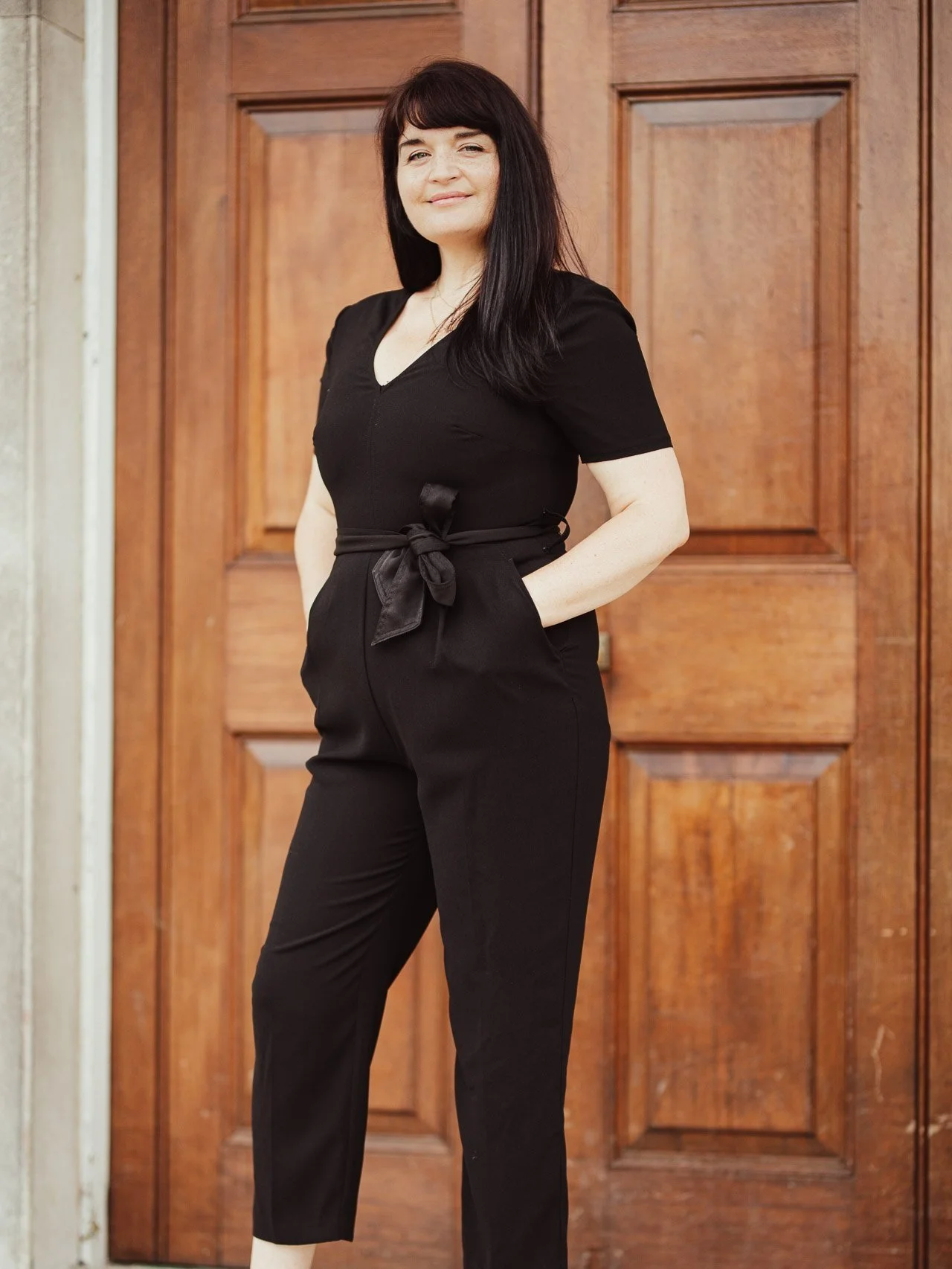 Woman (Monique Shaw) with long dark hair wearing a black outfit standing in front of a wooden door.