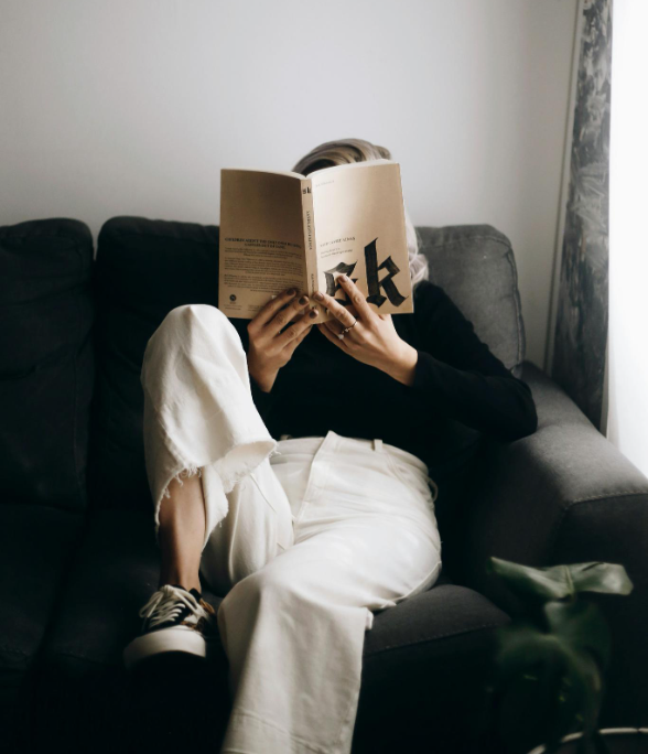 Person relaxing on a dark gray sofa, reading a book, with one leg crossed over the other, near a window with curtains.