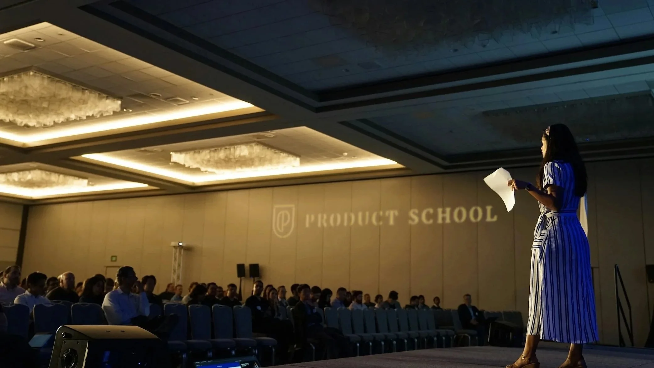 A woman standing on stage holding a paper, addressing an audience at a conference in a large, decorated conference hall with chandeliers and a sign that reads 'Product School.'
