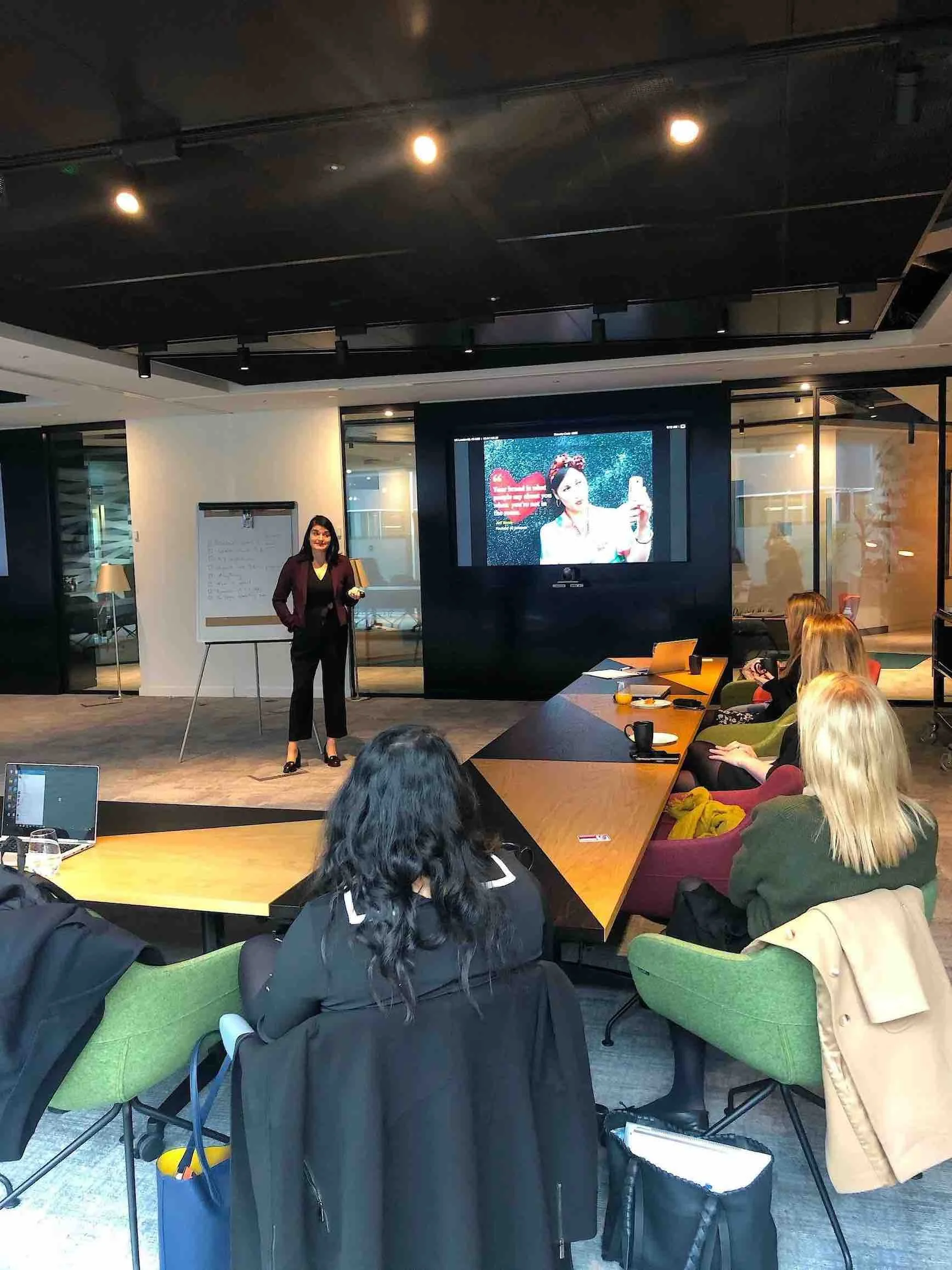 A woman in a dark blazer and black pants stands in front of a group, presenting in a conference room with a large screen displaying a woman taking a selfie with a heart graphic. Attendees are sitting around a U-shaped table, some taking notes or photos.