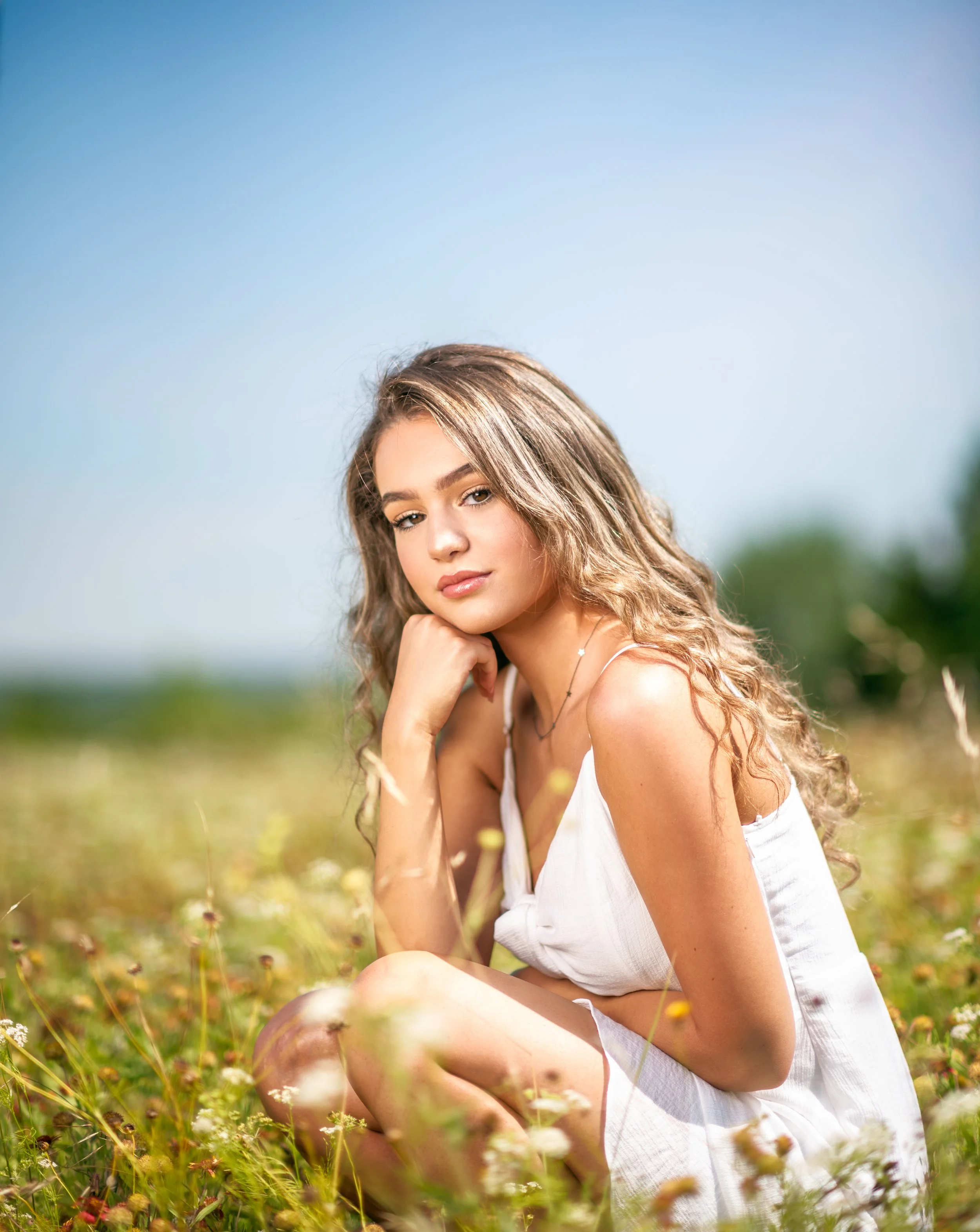 Woman in white dress sitting in a flower-filled meadow under a clear blue sky.