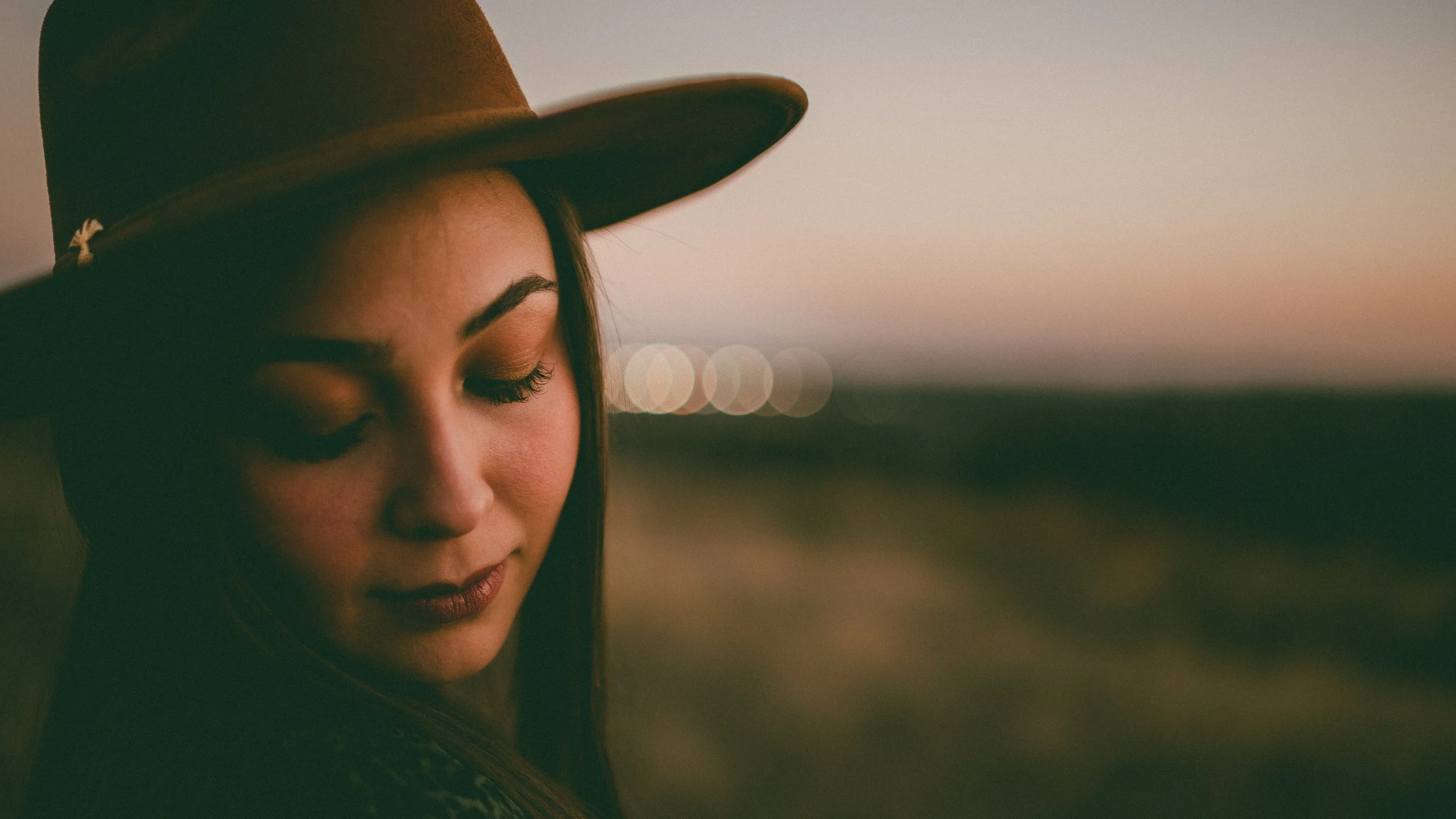 Close-up of a woman wearing a wide-brimmed hat, looking down with closed eyes, against a blurred outdoor background during sunset.