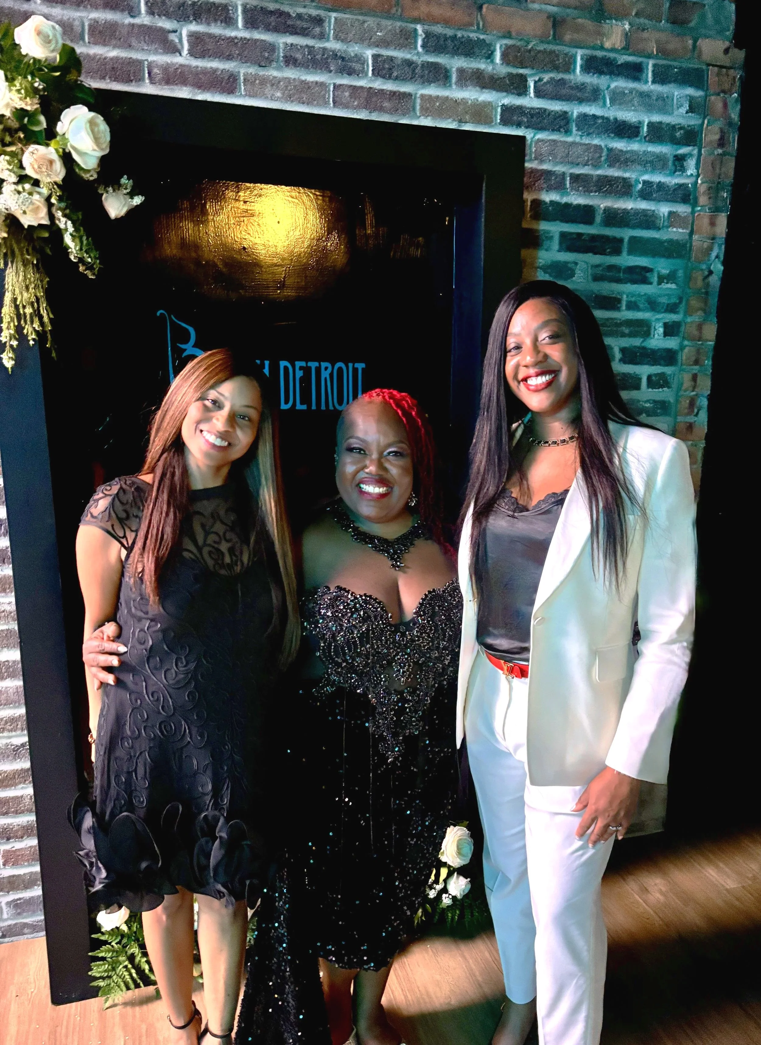 Three women smiling in front of a brick wall with a floral arrangement on the left. The woman in the center is wearing a black, sequined gown with beadwork. The woman on the right is wearing a white suit with a black top. The woman on the left is wearing a black dress with patterned detailing.