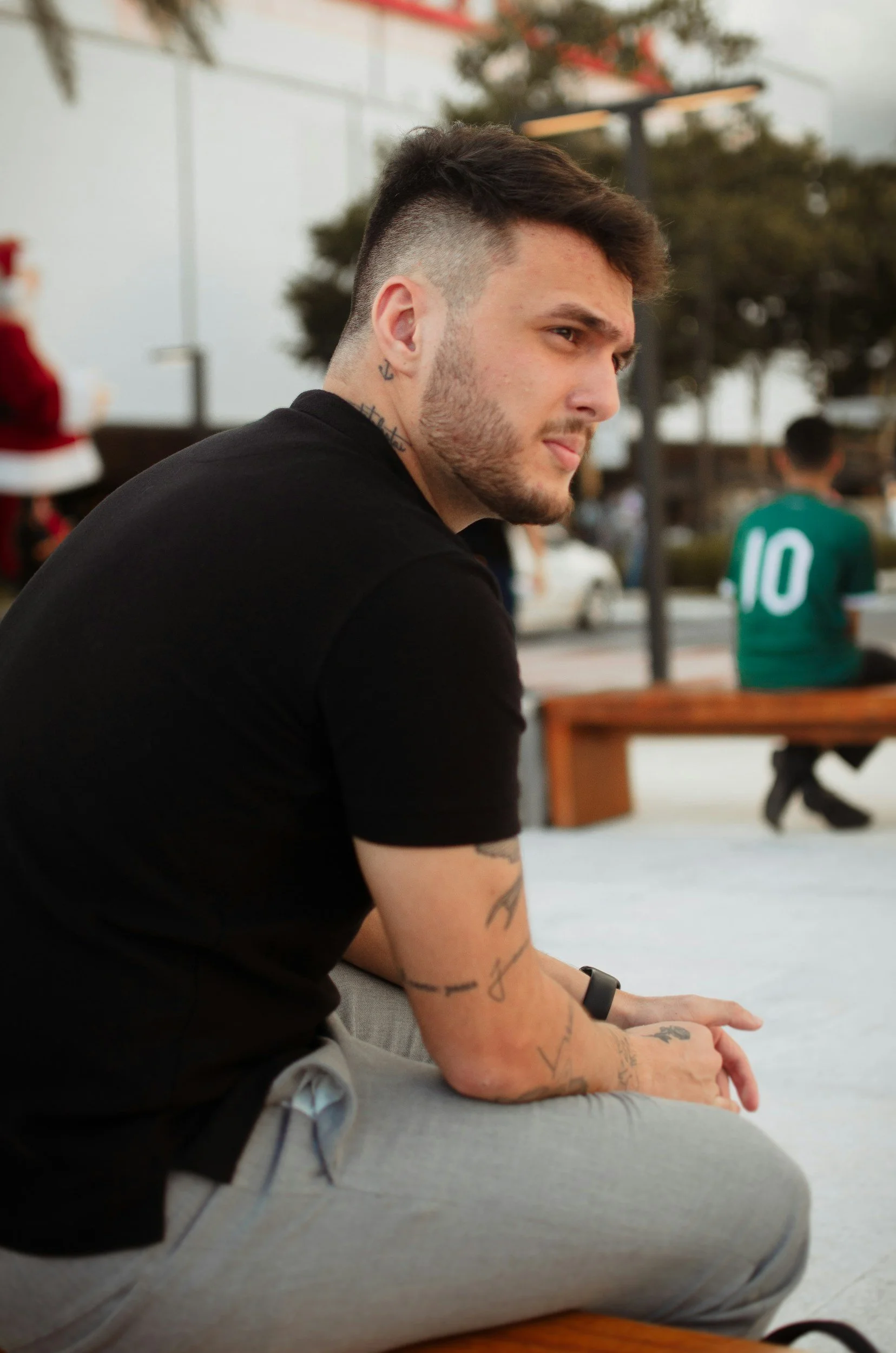 A young man with tattoos on his arm and neck, sitting outdoors with a contemplative expression, wearing a black t-shirt and gray pants.