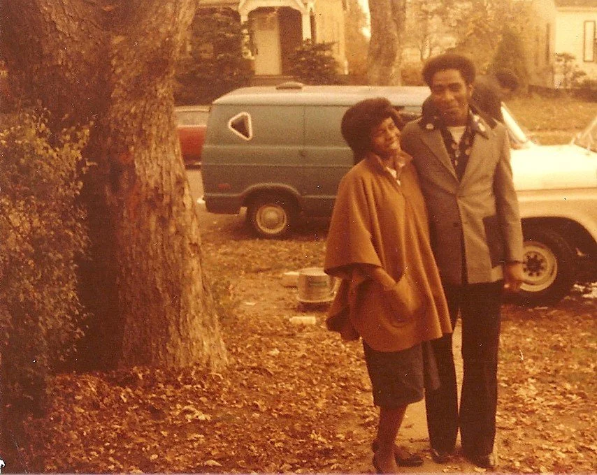 A vintage photograph shows an African American woman and a tall man standing outdoors next to a tree, with cars in the background during fall.