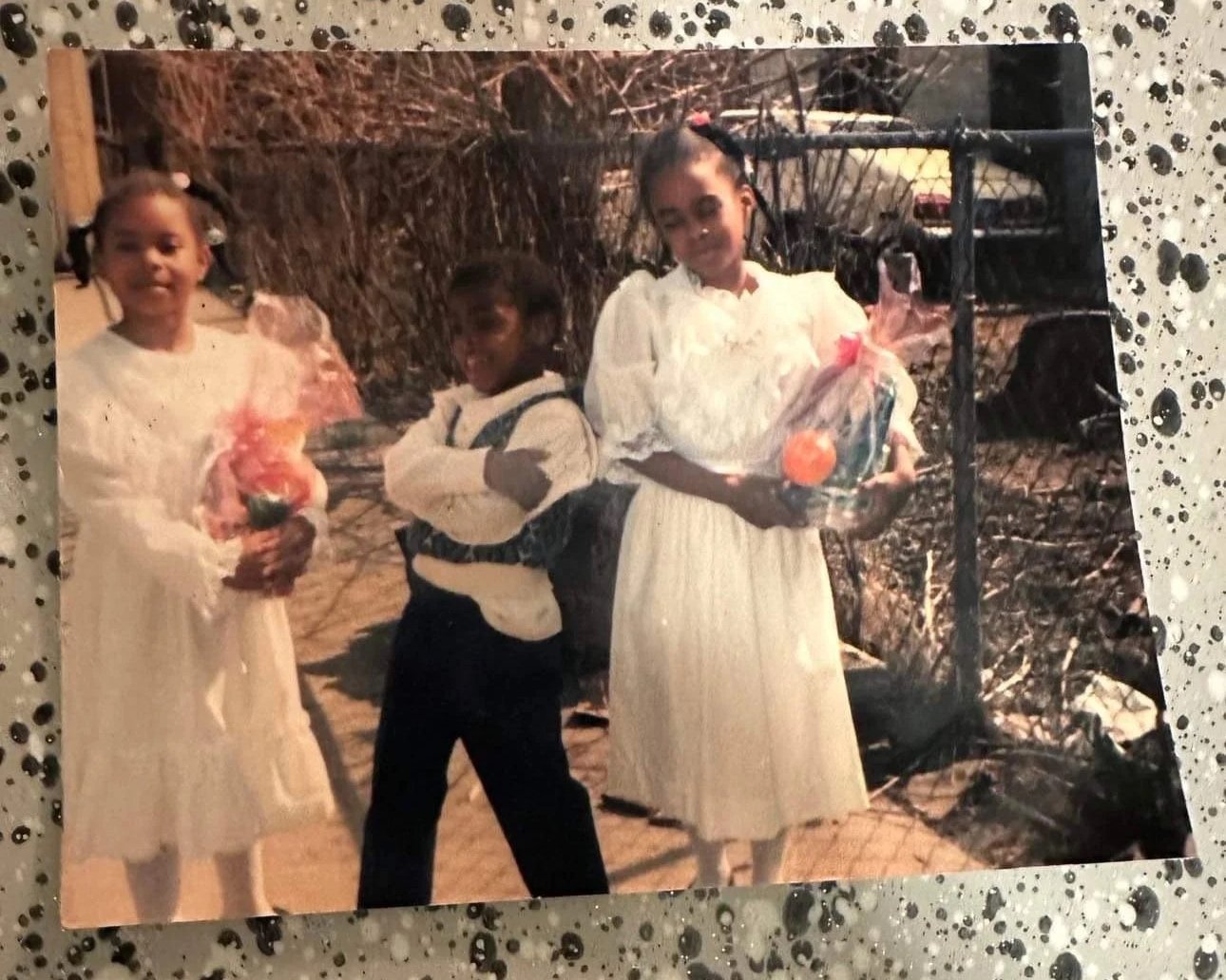 Three children standing outdoors, holding gift bags, with a tire and trees in the background.